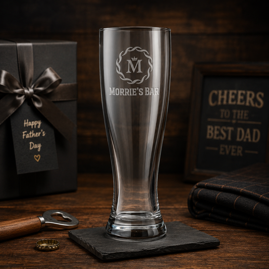 Clear glass with engraved text on a dark wooden surface with a gift and sign in the background.