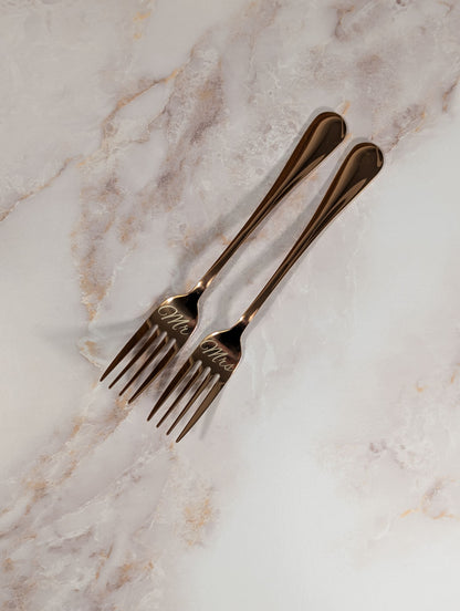 Two rose gold forks with 'Mr' and 'Mrs' engraving on a marble surface